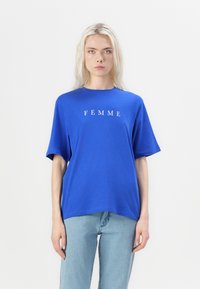 Blauwe katoenen T-shirt met korte mouwen, ronde halslijn en de tekst "FEMME" in het wit gedrukt over de borst. G gedragen met lichtblauwe jeans.