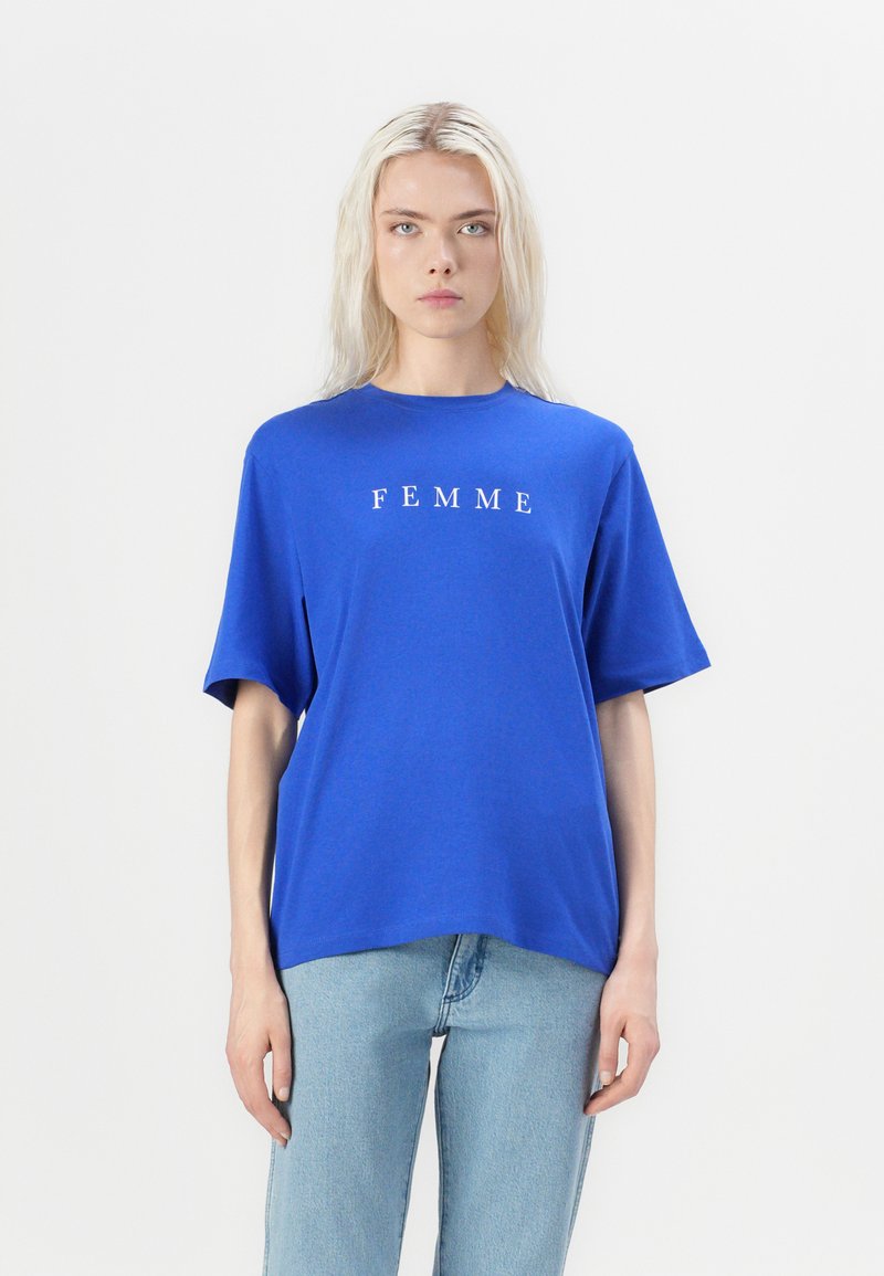Blauwe katoenen T-shirt met korte mouwen, ronde halslijn en de tekst "FEMME" in het wit gedrukt over de borst. G gedragen met lichtblauwe jeans.