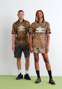 Gepardo rašto trumpomis rankovėmis marškiniai su žaliais akcentais ir Umbro logotipu. Nešiojami su pilkomis šortais ir sijonais, derinami su juodais aksesuarais.