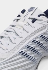 Κοντινό πλάνο λευκού αθλητικού παπουτσιού με πάνω μέρος από δίχτυ, κορδόνια και μαύρες ρίγες στη σόλα, με την επωνυμία "Hypercourt Express 3."