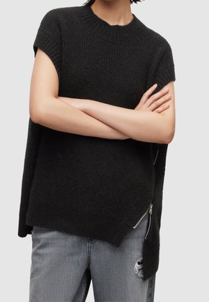 Persoon draagt een zwart mouwloos gebreid vest met een ritssluiting aan de zijkant en lichtgrijze ripped jeans, armen gekruist over de borst.