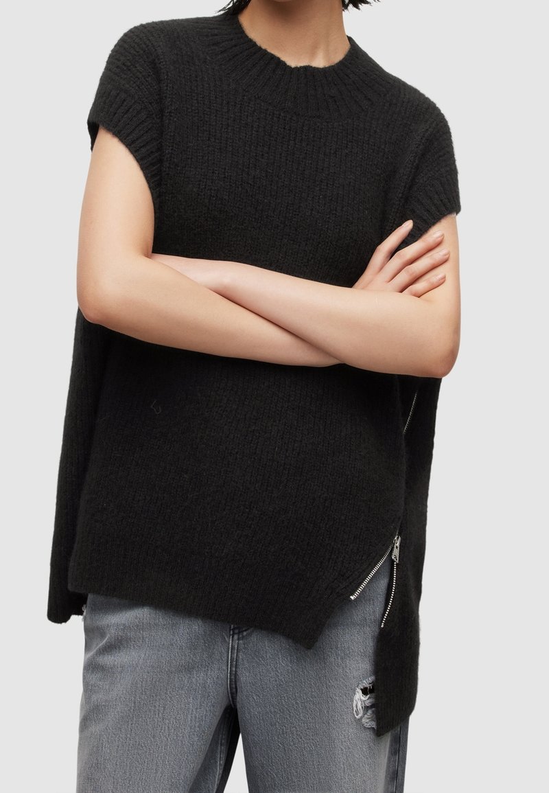 Personne portant un pull sans manches en maille noire avec un détail de fermeture éclair sur le côté et un jean gris clair déchiré, les bras croisés sur la poitrine.