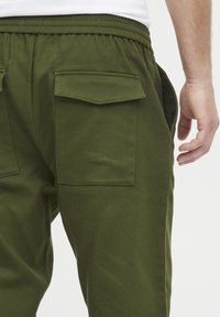 Olivenfargede joggebukser med elastisk midjebånd, sidelommer og to baklommer med knappeklapper. Laget av myk bomullsblanding.