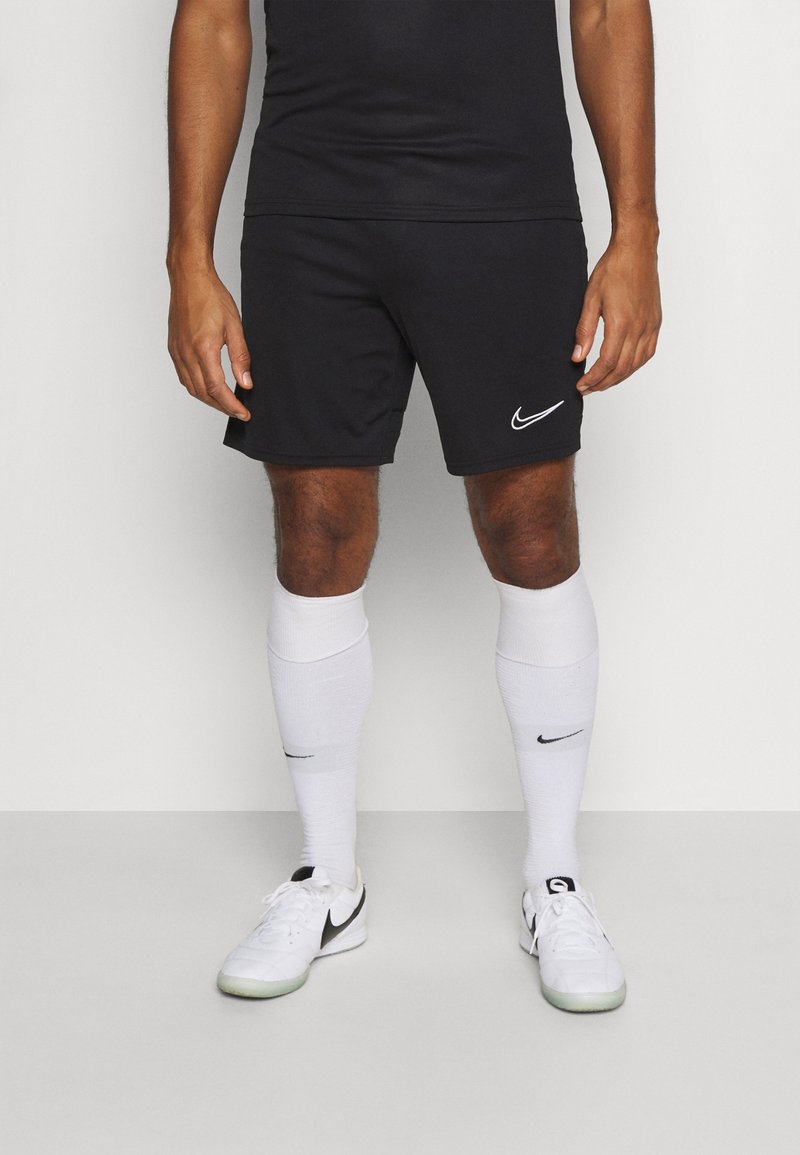 Homme portant un short Nike noir, des chaussettes blanches Nike montantes jusqu'aux genoux et des chaussures de sport blanches Nike, debout devant un fond uni.