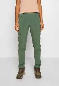 Groene wandelbroek met een taps toelopende pasvorm, elastische tailleband en zijzakken, gecombineerd met grijze en bruine wandelschoenen.