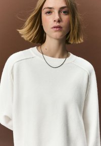 Biely sveter s voľným strihom a žebrovaným výstrihom, vyznačujúci sa jemnou textúrou. Na krku je tenký reťazový náhrdelník.