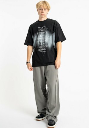 Młoda osoba o blond włosach, ubrana w czarną koszulkę z nadrukiem, luźne szare spodnie i czarne buty, stojąca na białym tle.