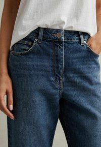 Kvinde iført mørkeblå jeans med mellem talje og en hvid tucket t-shirt, med den ene hånd i lommen og den anden afslappet ved siden.