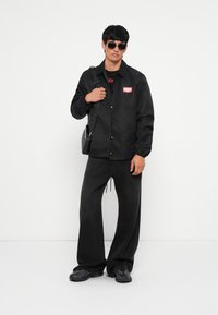 Μαύρο ελαφρύ μπουφάν με λογότυπο, σε συνδυασμό με άνετα μαύρα τζιν παντελόνια και μαύρα αθλητικά παπούτσια. Τα αξεσουάρ περιλαμβάνουν μια τσάντα χιαστί και γυαλιά ηλίου.