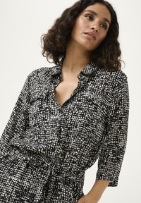 Camisa de botones en blanco y negro con un patrón abstracto texturizado. Presenta un cuello, dos bolsillos frontales y una cintura ajustable con lazo.
