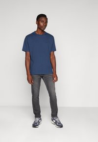 Maglietta in cotone blu navy con scollatura a girocollo, abbinata a jeans grigi e sneakers con motivo. La maglietta presenta un marchio discreto sul petto.