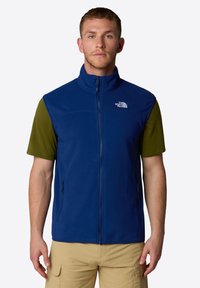 Gilet in pile blu con colletto alto, chiusura a zip e due tasche laterali. Indossato sopra una maglietta verde a maniche corte. Presenta un logo sul lato sinistro del petto.