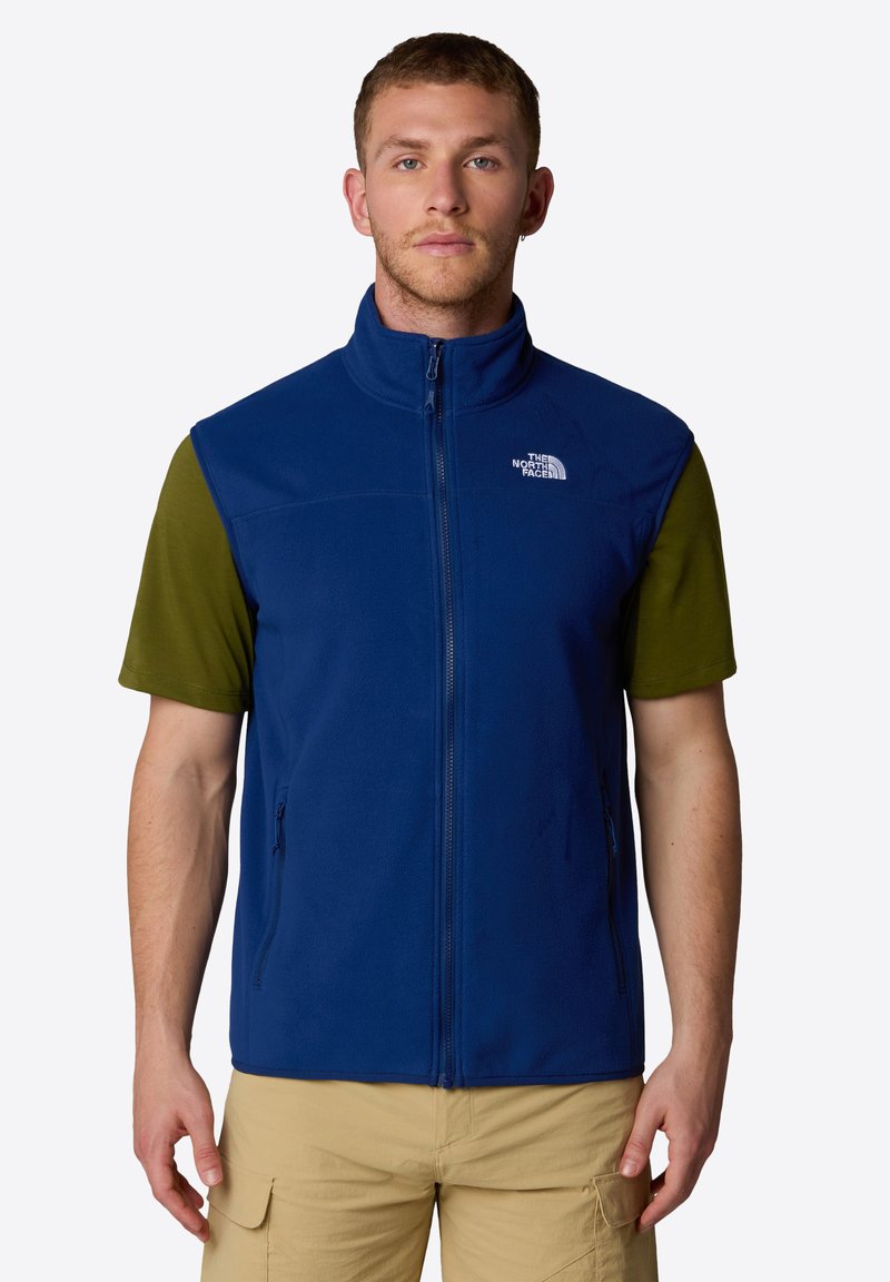 Gilet in pile blu con colletto alto, chiusura a zip e due tasche laterali. Indossato sopra una maglietta verde a maniche corte. Presenta un logo sul lato sinistro del petto.