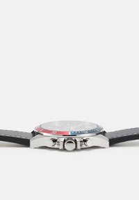 Reloj de acero inoxidable plateado con correa de cuero negro, bisel rojo y azul, y tres botones en el lado para ajustes.