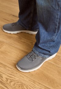 Grå nät sneakers med vit sula, med elastiska snören och en strukturerad ovandel, i kombination med blå denimjeans på ett trägolv.