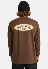 Camisa de manga larga marrón con un gran logo amarillo y blanco en la parte posterior que presenta un gráfico de ola estilizado. Material de algodón, corte relajado.
