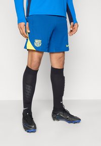 Μπλε αθλητικό σορτς με κίτρινες λεπτομέρειες και λογότυπο συλλόγου, συνδυασμένο με μαύρες κάλτσες έως το γόνατο και μαύρα παπούτσια με μπλε λεπτομέρειες.