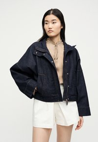 Γυναίκα με μακριά σκουρόχρωμα μαλλιά φορά υπερμεγέθη σκούρο τζιν μπουφάν πάνω από μπεζ μπλούζα και λευκά σορτς, στέκεται μπροστά σε απλό φόντο.