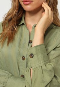 Camisa verde oliva con rayas verticales, que presenta grandes botones marrones y puños fruncidos, hecha de una tela ligera y suave.