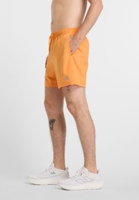 Oranssit uima-shortsit elastisella vyötäröllä ja nyörillä, joissa on sivutaskut ja pieni harmaa logo, yhdistettynä valkoisiin kenkiin.