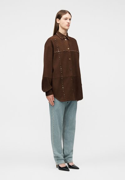 Γυναίκα που φοράει υπερμεγέθη καφέ πουκάμισο με κουμπιά και τρουκς, ανοιχτού μπλε τζιν παντελόνι με ίσια γραμμή και μαύρες μυτερές γόβες, στέκεται μπροστά σε απλό φόντο.