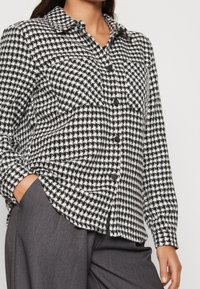 Camicia a scacchi bianco e nero con colletto, maniche lunghe e bottoni neri, abbinata a pantaloni grigi. Tessuto testurizzato con un sottile motivo.