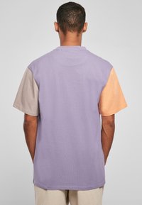 Triko s krátkým rukávem v levandulové barvě, béžovým levým rukávem a broskvovým pravým rukávem; vyrobené z bavlny s volným střihem.