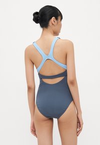 Maillot de bain une pièce avec un corps gris foncé et des bretelles bleu clair. Le design inclut un dos ouvert et des découpes.