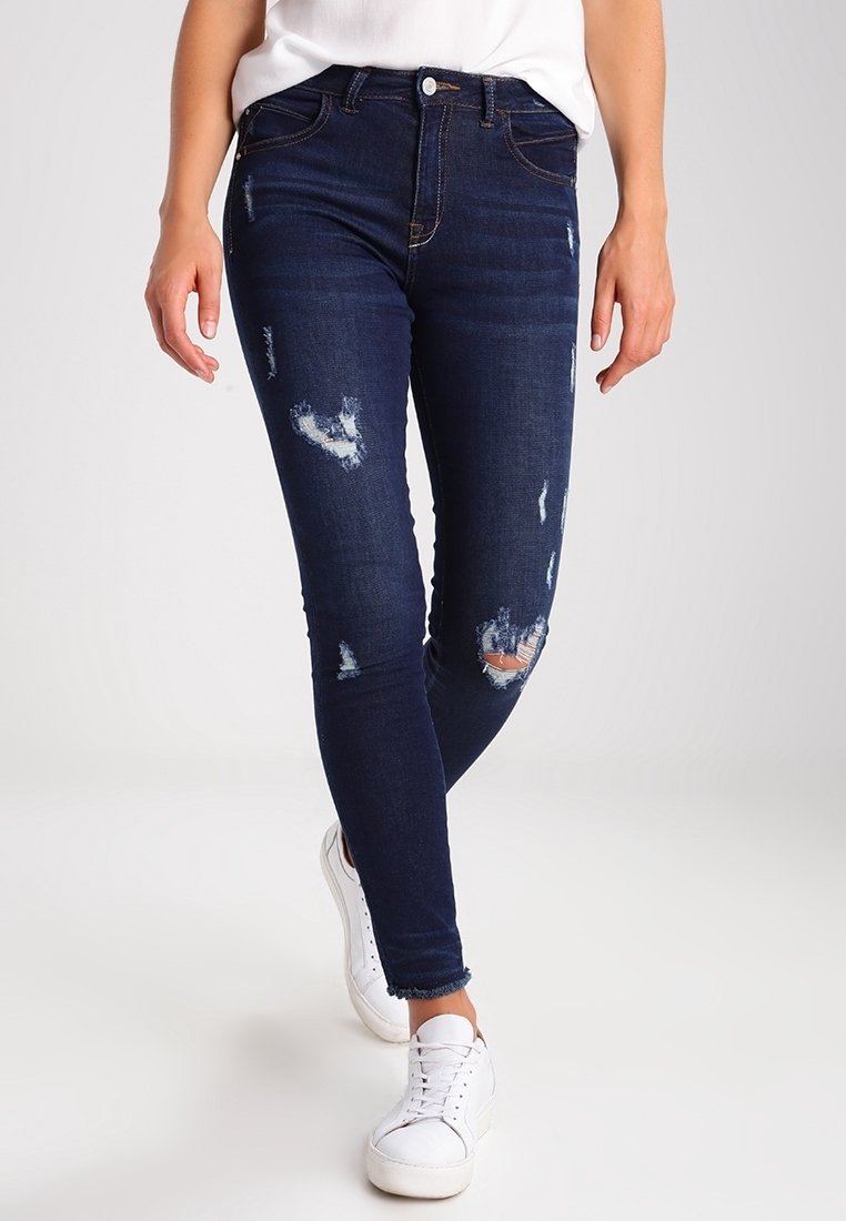 Jeans ajustados de un azul oscuro con parches desgastados, que presentan un diseño clásico de cinco bolsillos y un ligero desgaste. Combinados con zapatillas blancas.