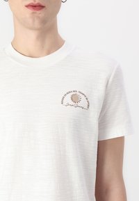 Hvit t-skjorte med korte ermer laget av teksturert stoff. Har et lite brunt grafisk design med tekst på venstre brystområde.