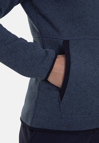 Fleecejack in donkerblauw met een gestructureerde stof. Heeft een verborgen zak aan de zijkant en contrasterend donker rand langs de randen.