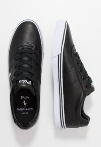 Svarta lädersneakers med rund tå, svarta snören och vit sula. Har logotyp på tungan och kontrasterande sidoflikar. Slät textur.