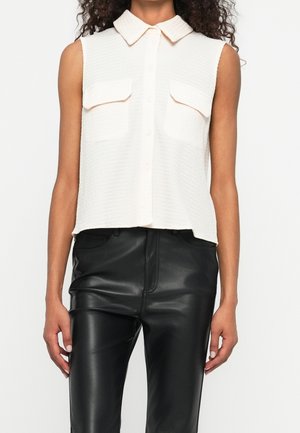 Kobieta ubrana w białą, bezrękawną koszulę na guziki z dwoma kieszeniami na piersi oraz czarne, skórzane spodnie z wysokim stanem, stojąca na tle białego tła.