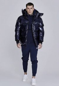 Mornarsko modra puhasta jakna z bleščečim zaključkom, z zadrgo in kapuco; kombinirana z mornarsko modro kapucarico in trenirkami, dopolnjena z belimi supergami.