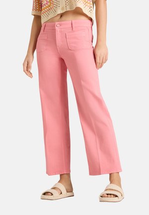 Vrouw met een hooggerokte, wijdere roze broek, een cropped gehaakt top en beige gevlochten instapsandalen, staand op een witte achtergrond.