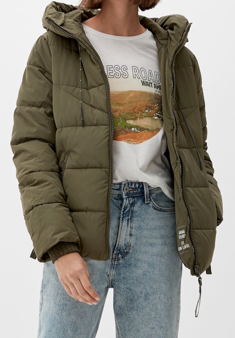 Olive green puffer jacket with a hood, featuring a zip closure, elastic cuffs, and a logo patch on the side, worn over a graphic tee.