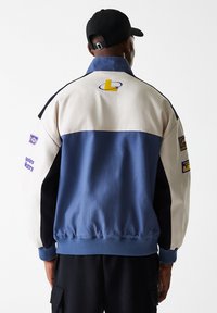 Blue, cream, and black color-blocked jacket with ribbed cuffs, featuring Los Angeles Lakers logos on sleeves and back. Smooth fabric texture.