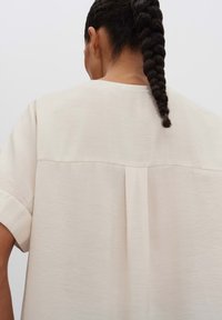 Camicia beige chiaro con finitura texturizzata, maniche corte arrotolate e dettaglio a pieghe sul retro per un tocco di design in più.