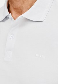 Biała koszulka polo z teksturowanej tkaniny, z zapięciem na guziki u kołnierzyka, małym haftowanym logo "P.L." na piersi oraz subtelnym detalem w przeszyciach.