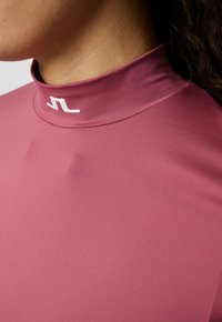 Mauve högkragsidrottstopp gjord av ett slätt, elastiskt material med en subtil glans, med en vit logotyp vid kragen.