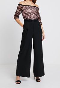 Combinaison large noire avec un buste en dentelle florale rose et noire décolleté, présentant des manches trois-quarts et une taille ajustée.