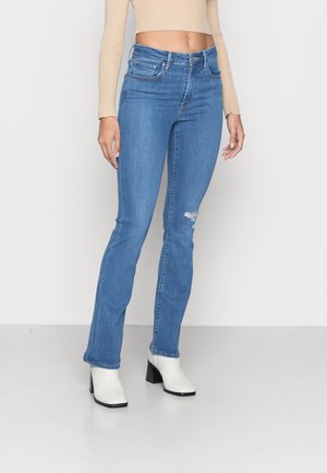 Kvinde iført lyseblå højttaljede skinny jeans med let slid på knæet og hvide hælede ankelstøvler, beige cropped top synlig.