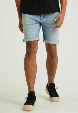 Hellblaue Denim-Shorts mit umgeschlagenen Säumen, die Verwitterungen und fransige Details aufweisen, kombiniert mit schwarzen Sneakers mit beigefarbenen Sohlen.
