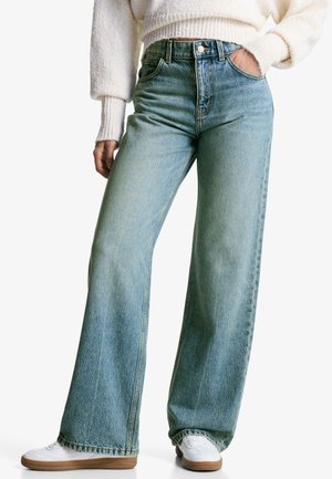 Person iført højtaljede, vide blå jeans, hvide sneakers og en cremefarvet striktrøje med den ene hånd i lommen.