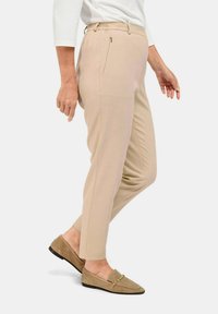 Beige schmal zulaufende Hosen mit einem seitlichen Reißverschluss und elastischem Bund, kombiniert mit hellbraunen loafers mit goldenen Akzenten.