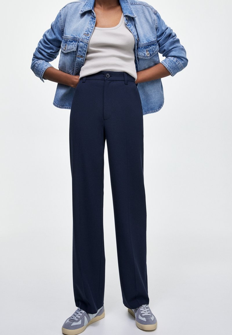 Personne portant une veste en jean bleu clair par-dessus un débardeur blanc, un pantalon large bleu marine et des baskets grises, debout les mains sur les hanches.