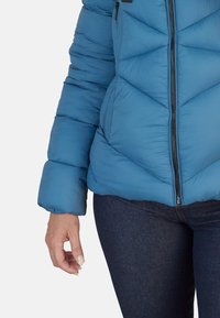 Chaqueta acolchada azul con costuras en forma de chevrón, cierre de cremallera y cuello alto, combinada con jeans oscuros. Presenta un acabado ligeramente texturizado.
