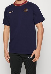 Camiseta de manga corta azul marino de algodón, con un cuello redondo color burdeos, logo dorado en el pecho y detalle del swoosh de Nike.