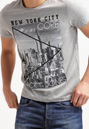 Homme portant un t-shirt gris avec un graphisme de la skyline de New York et un texte noir, assorti à un jean bleu, présenté de la nuque jusqu'aux hanches.