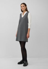 Vrouw in een grijs streepjesmouwloos jurkje over een witte blouse met lange mouwen, zwarte tights en zwarte instapschoenen, staande tegen een effen muur.
